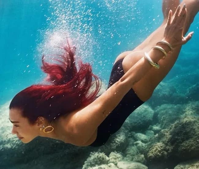 Dua Lipa takes a deep dive wearing a black one-piece swimsuit, with flowing auburn red hair on an alternative cover art for her 3rd studio album 'Radical Optimism'.