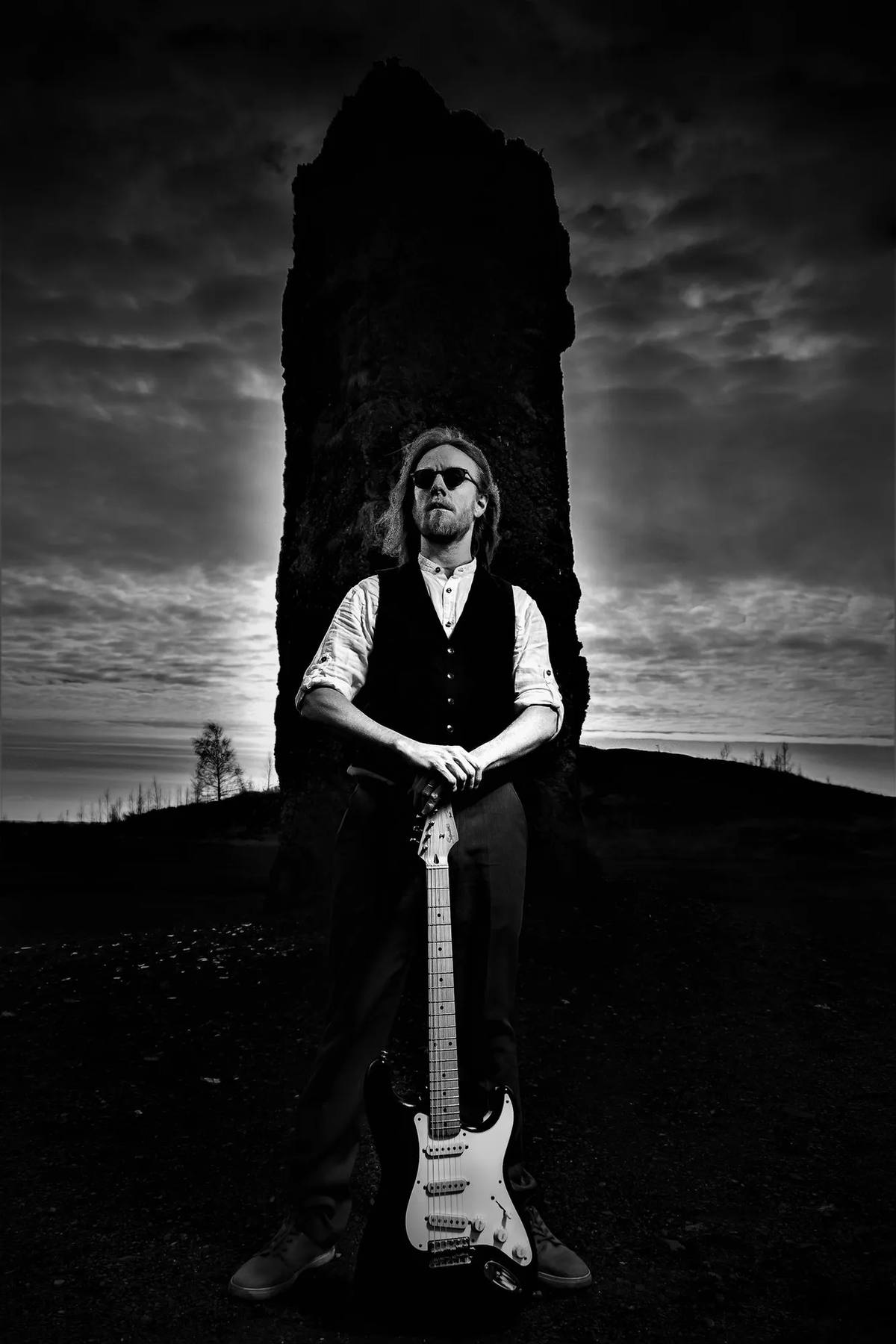Cannonball standing against large rock while holding onto his guitar, black and white.