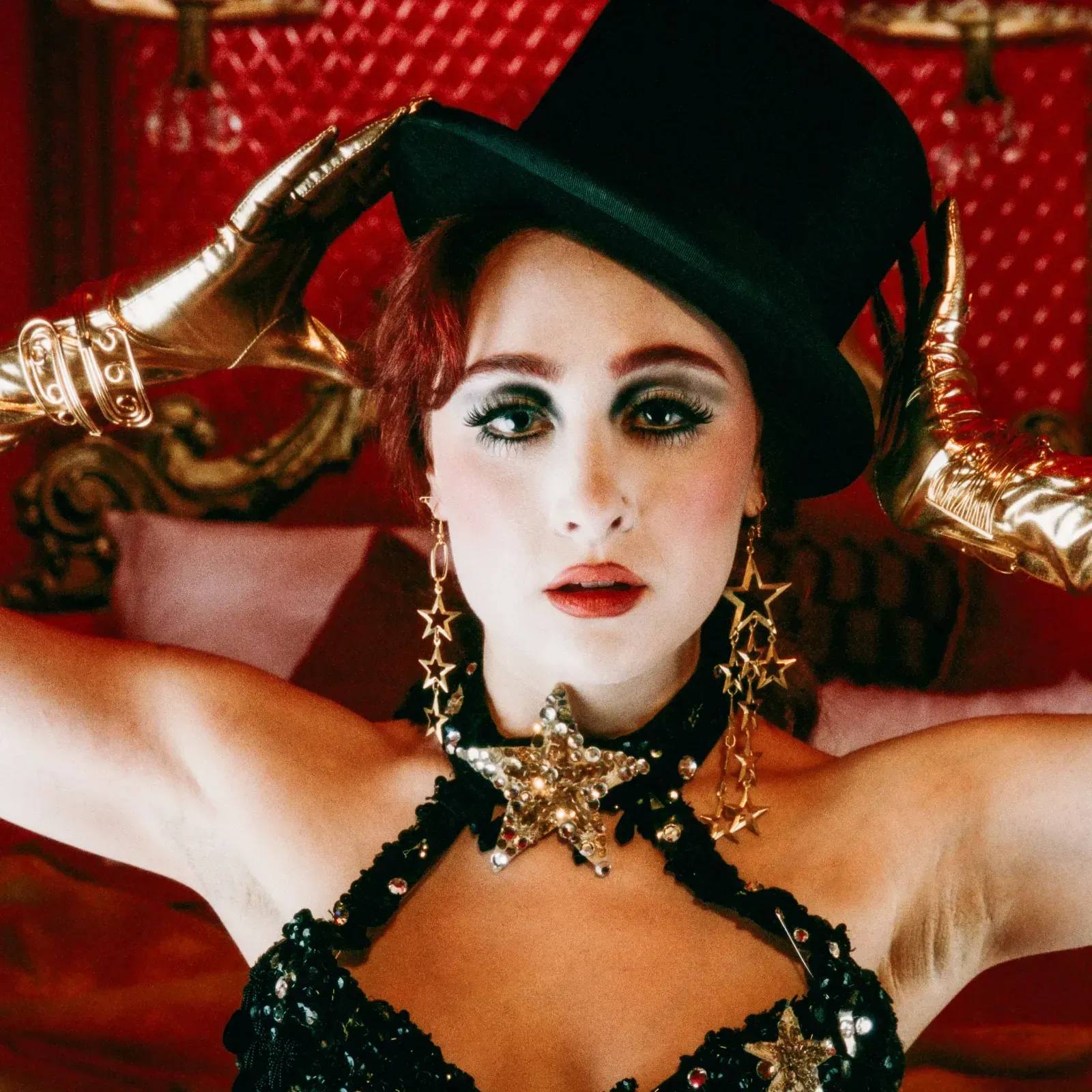 Chappell Roan poses for 'Red Wine Supernova' wearing a sparkling bra, green cylinder hat, and gold star earrings against a bright red background.