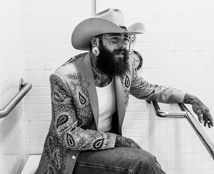Post Malone wears a country hat and an embellished jacket, holding a cigarette in a black-and-white shot taken in a shower.