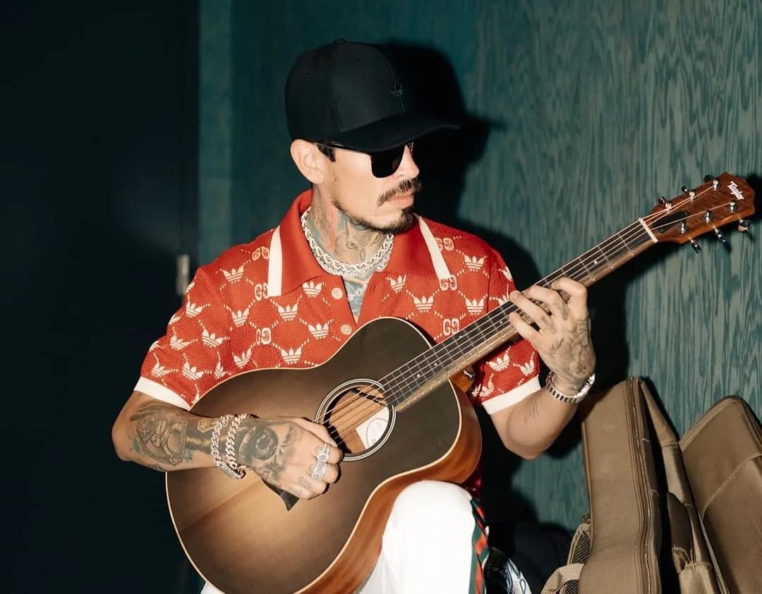 Codiciado, wearing a black Adidas hat, black sunglasses, an orange t-shirt, and white Gucci pants, plays an acoustic guitar. Golpes de la Vida Riaa gold.